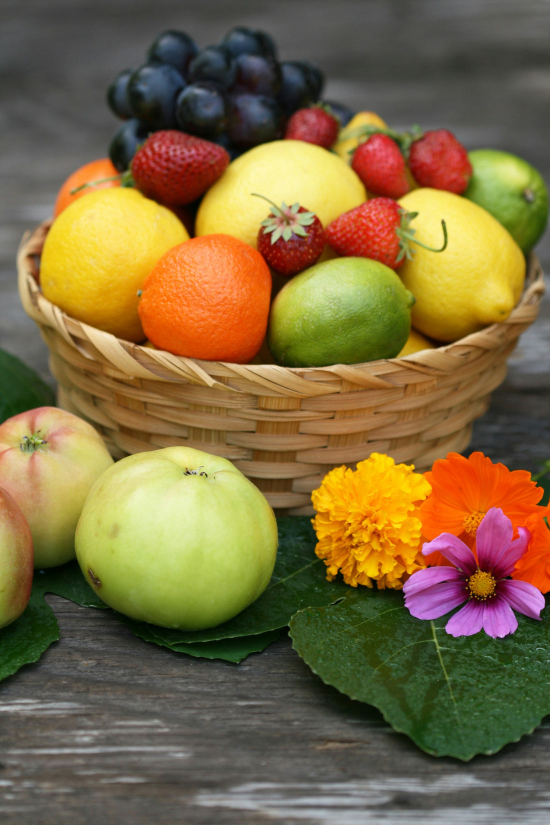 Panier en osier rempli de fruits frais : citrons, pommes, oranges, fraises, raisins et citrons verts, accompagné de fleurs colorées.